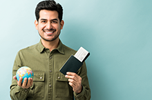 happy-young-man-holding-passport-boarding-pass-with-globe-while-standing-against-blue-background